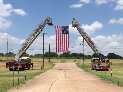 Image of two fire trucks displaying American flag from their ladders over a road
